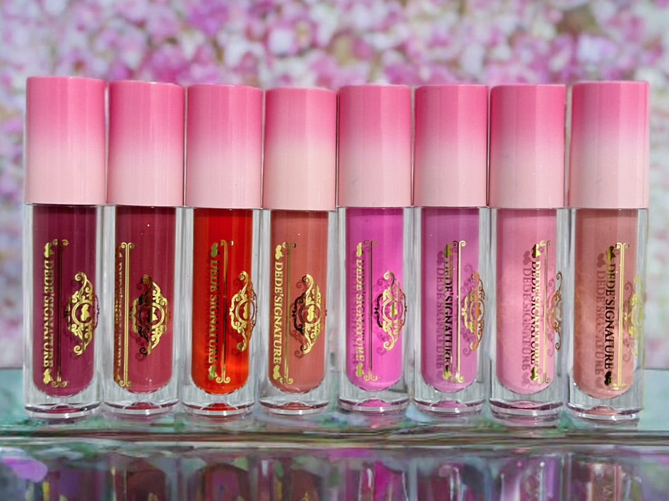 Eight Fleur & Blush liquid blush tubes with pink gradient caps lined up on a reflective glass surface, showing shades of pink, red, coral, and mauve, against a blurred pink floral background