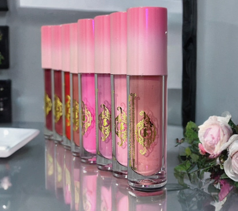 Six Fleur & Blush liquid blush tubes with pink gradient caps and gold ornamental labels arranged in a row on a reflective surface, with pink flowers at the right edge